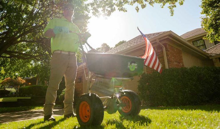 A picture from the ground of a man in a Green Group uniform standing in front of a green lawn with a fertilizer spreader