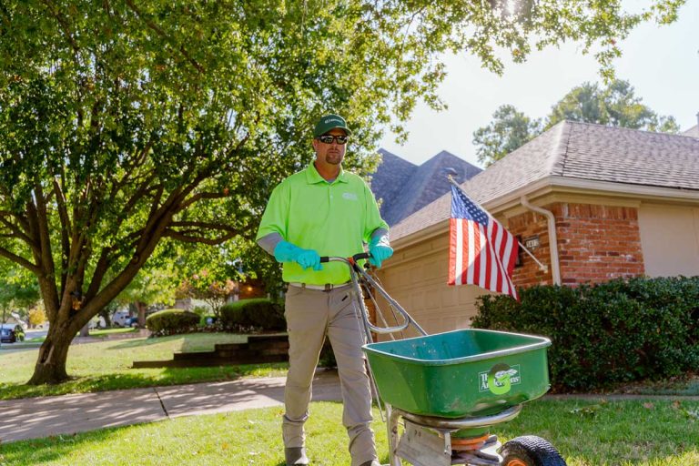 A man in a Green Group uniform standing in front of a green lawn with a fertilizer spreader